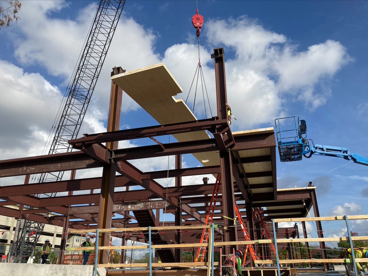 A construction project with a crane lifting items onto a steel structure. The sky is blue with white clouds.