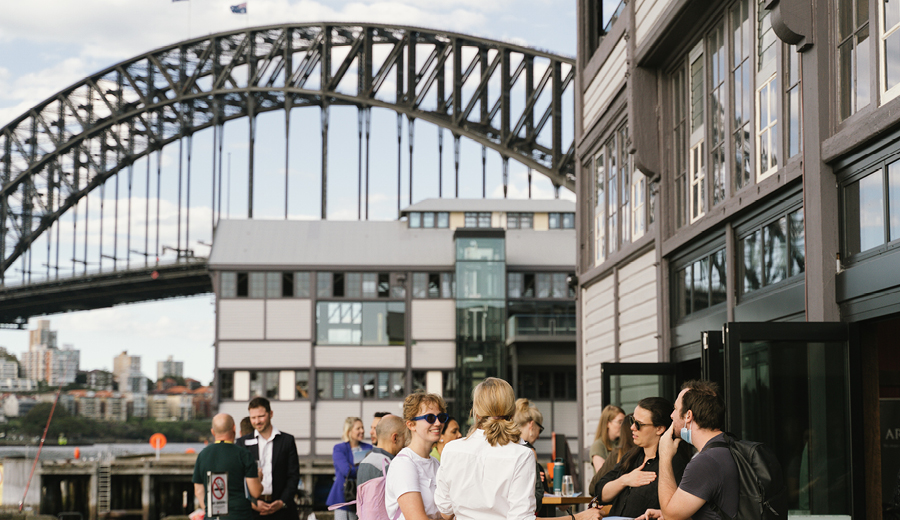 Outside at wharf in Sydney Harbour, a group of people are standing at tables and talking, day time