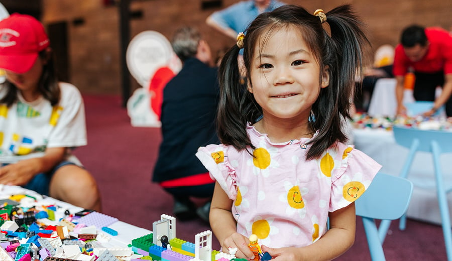 A happy young girl with straight black hair in pigtails looking at the camera, playing with bright coloured LEGO blocks 