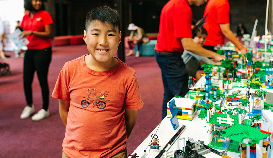 A boy smiling and wearing an orange t-shirt standing next to his creation of a city using LEGO blocks