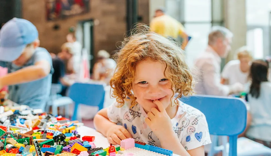 A young girl with curly red hair looking at the camera, playing with bright coloured LEGO blocks with a group of other children at an event