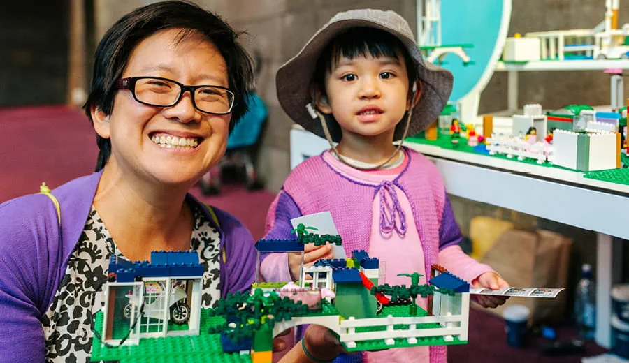 A woman with black, short hair and wearing glasses and very young child, both wearing pink and purple tops, are smiling and each holding a toy made out of coloured LEGO blocks