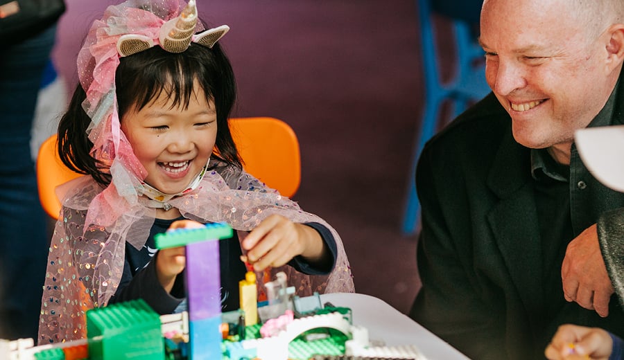 A young girl with black straight hair, wearing a pink coloured head-dress and cape resembling a unicorn is laughing and building a tower with LEGO blocks. A man in a black suit is smiling and looking on.
