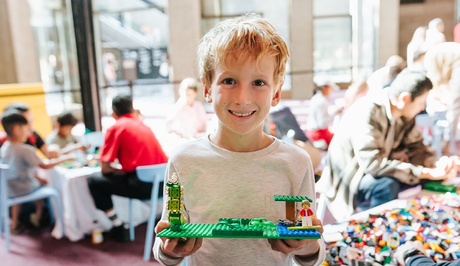A young boy with pale skin and blond hair smiles while holding his toy made out of LEGO blocks. 