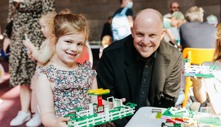 A young girl wearing a dress with printed flowers is smiling and holding a toy made out of LEGO blocks. A man in a black suit is smiling and looking on.