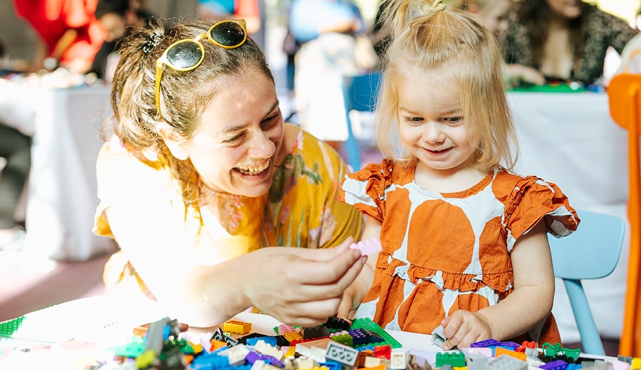 A woman smiling and wearing a yellow top is helping a very young girl, smiling and wearing a orange spotted dress, build a toy with LEGO blocks. 