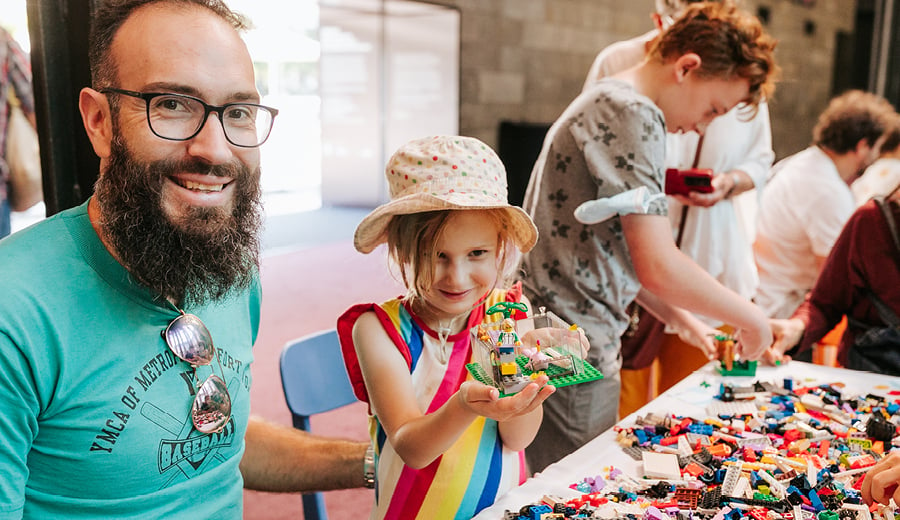 A man with a beard and glasses is sitting next to a young girl wearing a hat and brightly coloured striped dress. They are both happy. The girl is holding a toy made out of coloured LEGO blocks