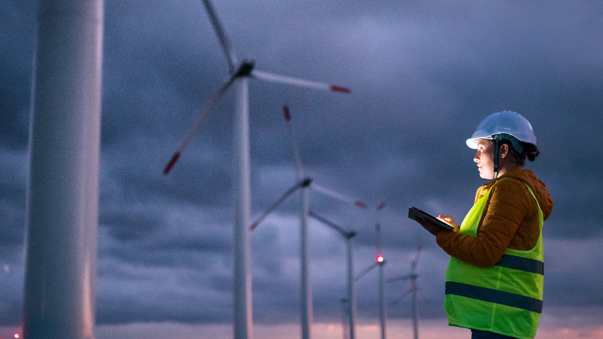 A femaile engineer holding a tablet looks over at a group of wind turbines and monitors them, at dusk.