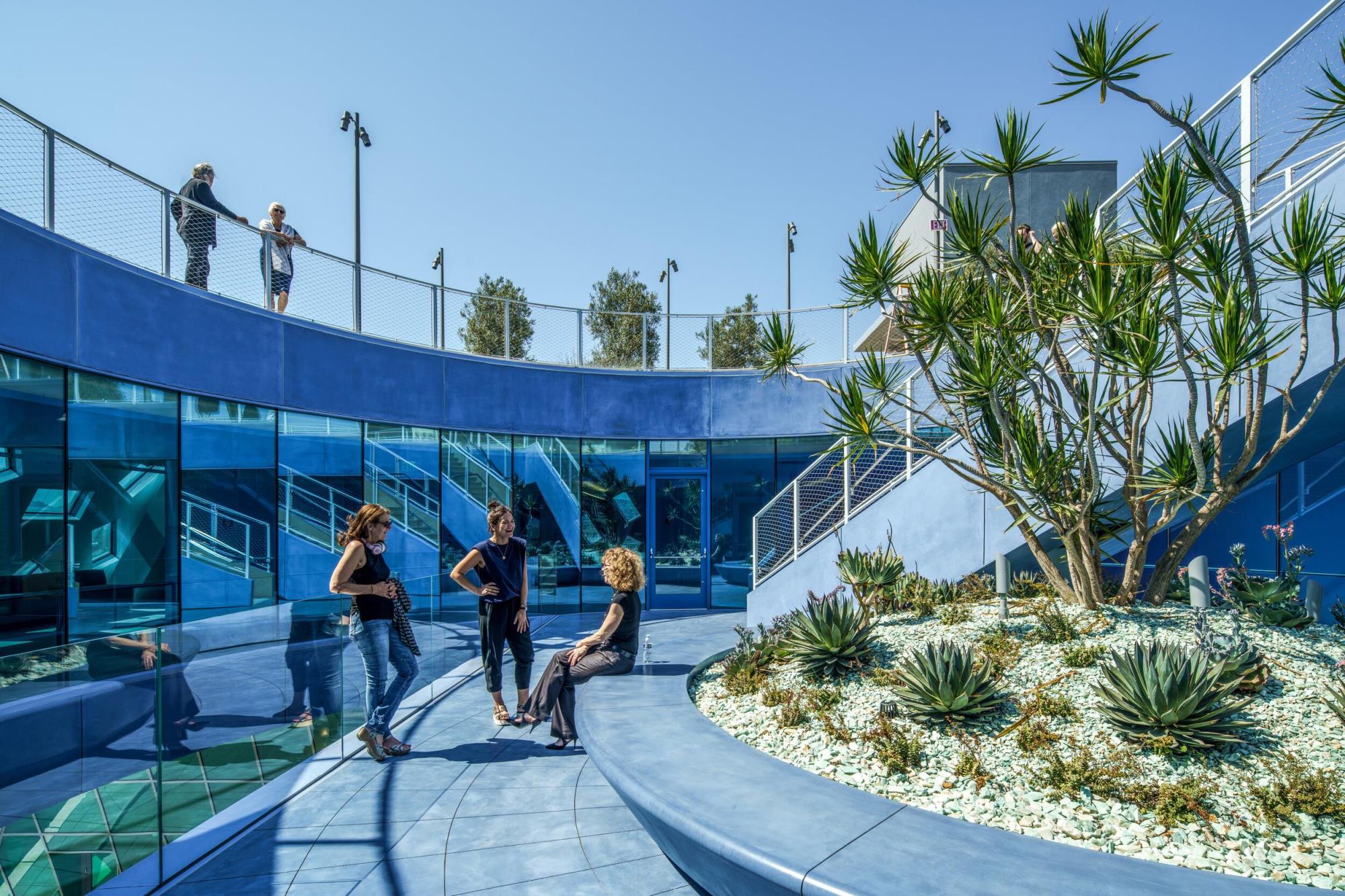Exterior view of the Audrey Irmas Pavilion courtyard. The space is comprised of blue and green glass as well as furniture. People are congregating.