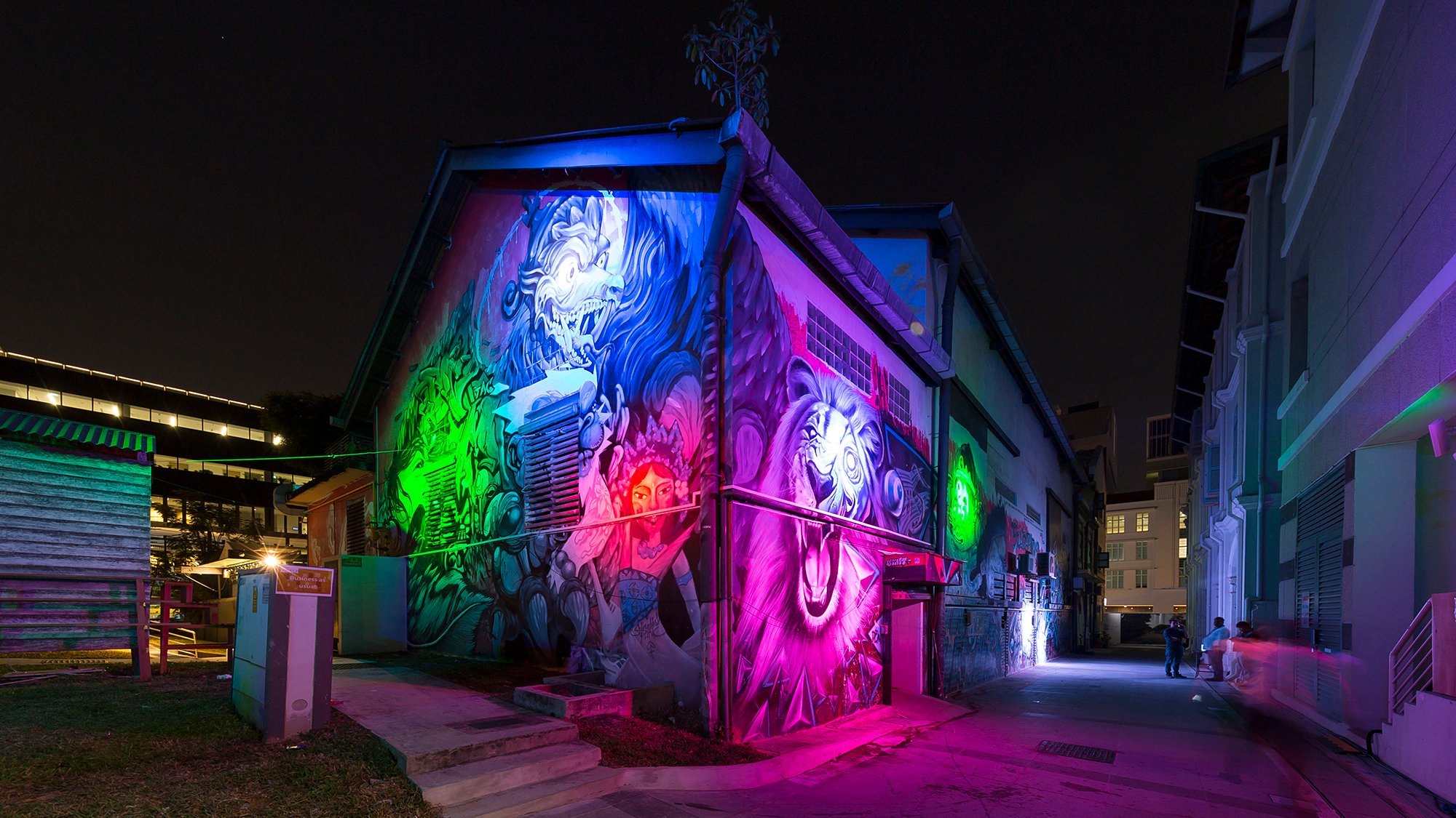 Night time photo of the corner of a building covered in graffiti and lit up by coloured spotlights