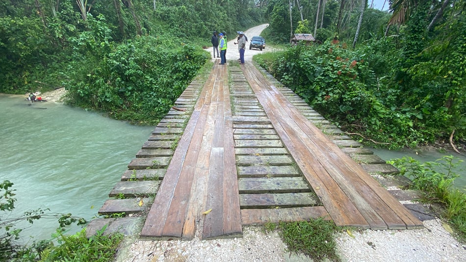 A view of a river running under a wooden bridge with green fiolage on the banks in a trropical location