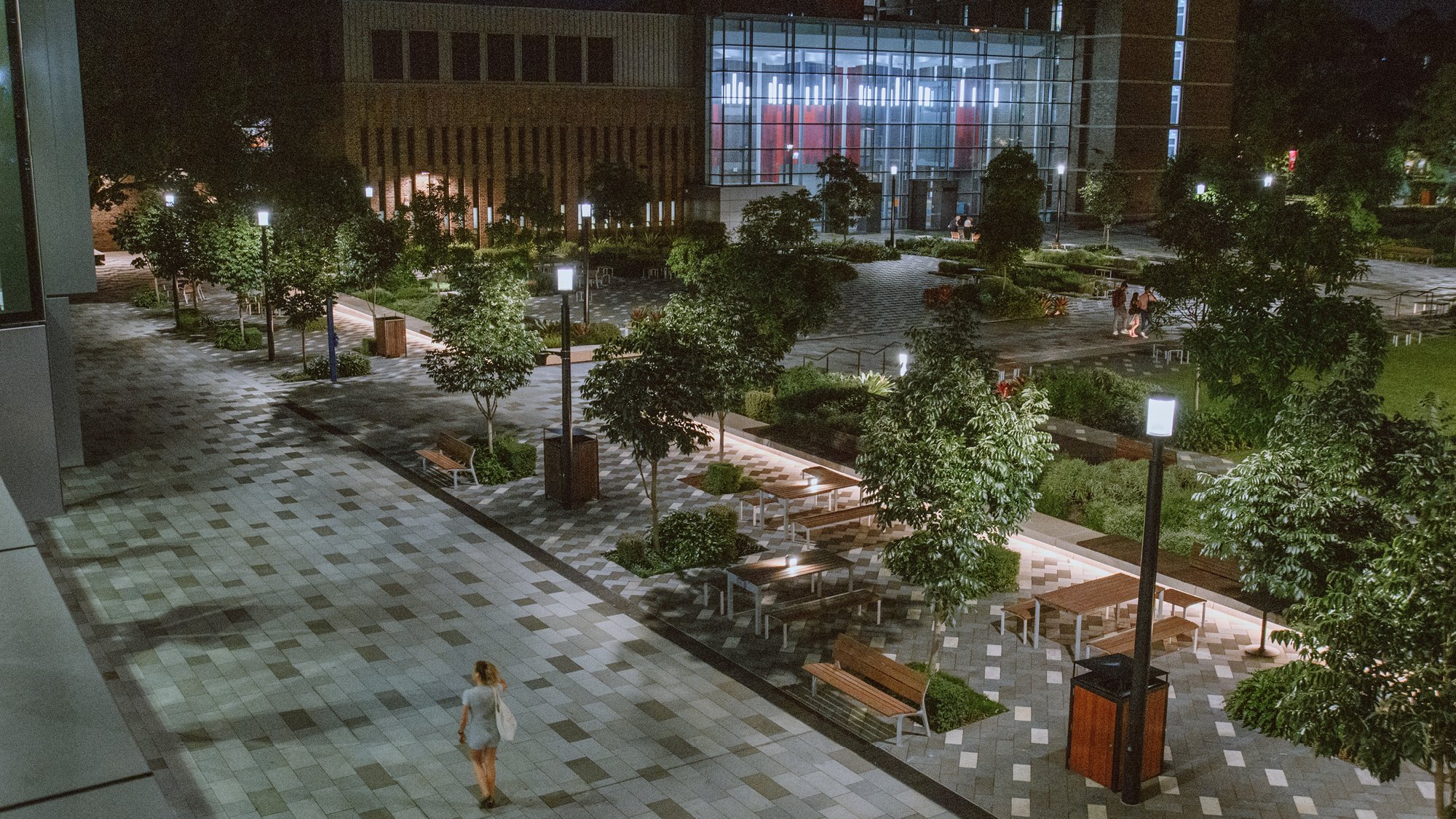 Woman walking through a large courtyard with trees and seating at night
