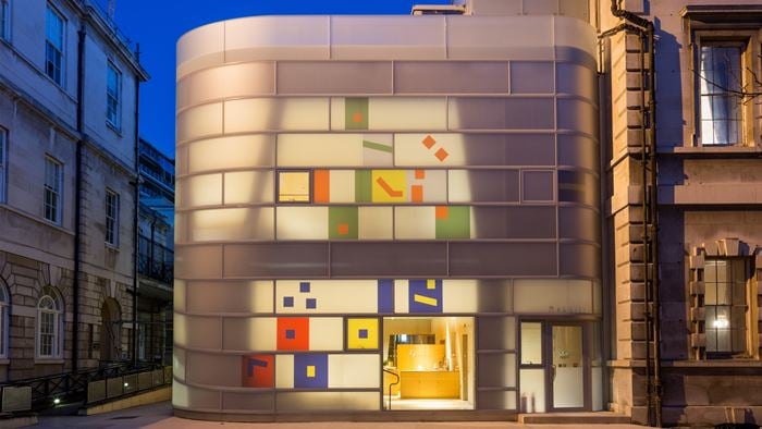 The curved, opaque glass building illuminated at night with sporadic coloured fragments on the façade.