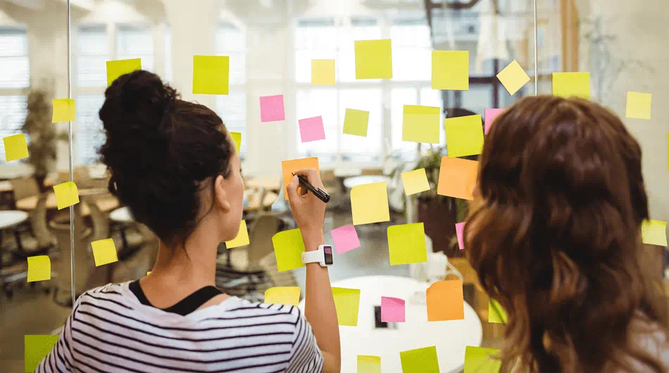 Two women writing on post it notes that are put on a transparent board