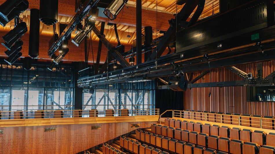 Inside a concert hall with folding seats, timber panels and spaces for orchestras. Hanging from the ceiling on rails is acoustic and lighting equipment 