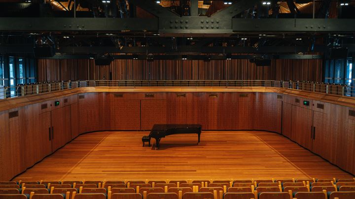 Inside a concert hall with grand piano, folding seats, timber panels and spaces for orchestras.