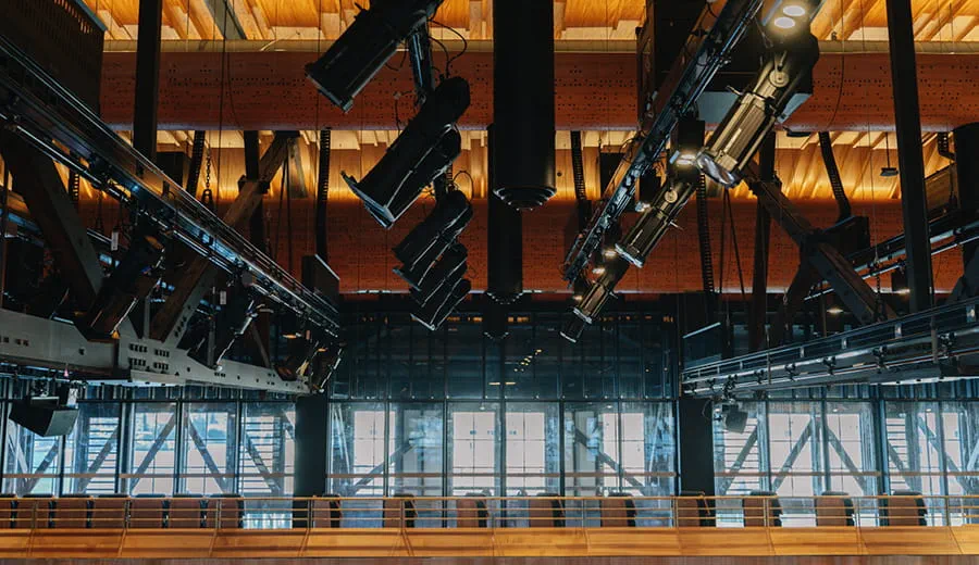 Inside a concert hall with folding seats, timber panels and spaces for orchestras. Hanging from the ceiling on rails is acoustic and lighting equipment