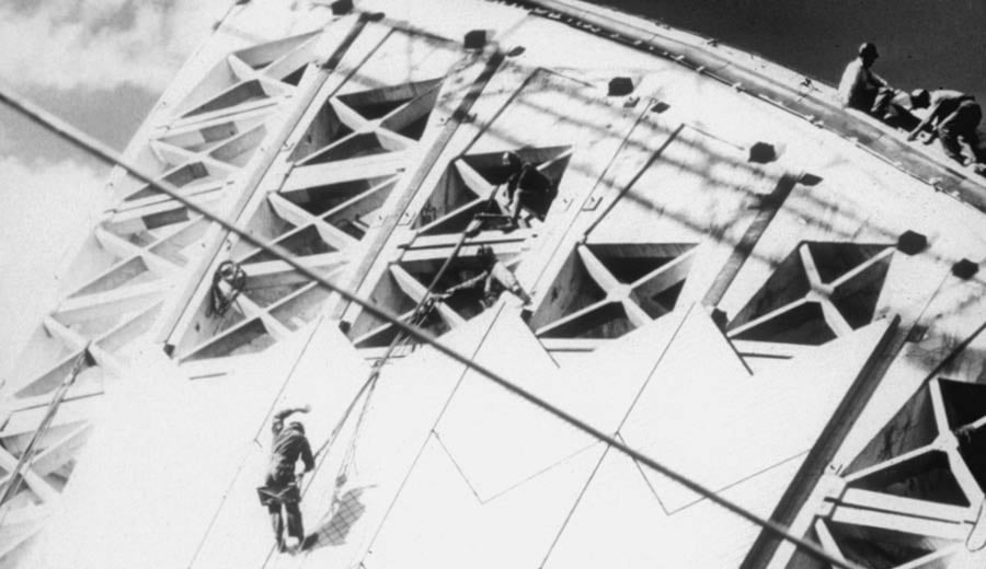 A black and white photograph of a man climbing on the roof sail at the Sydney Opera House during construction