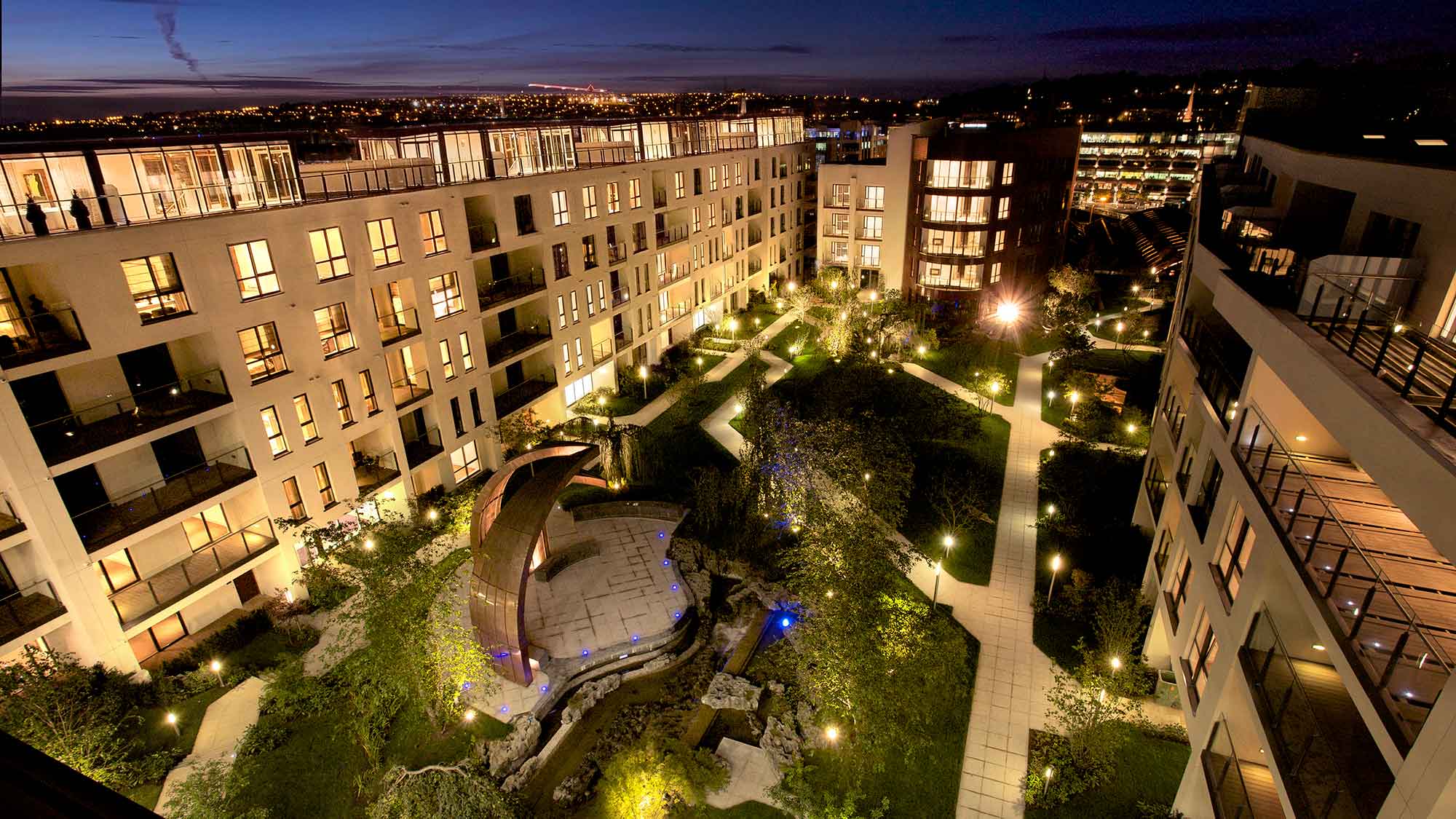 Night-time photo of apartments around courtyard garden, showing lighting around sculptures and on the paths.