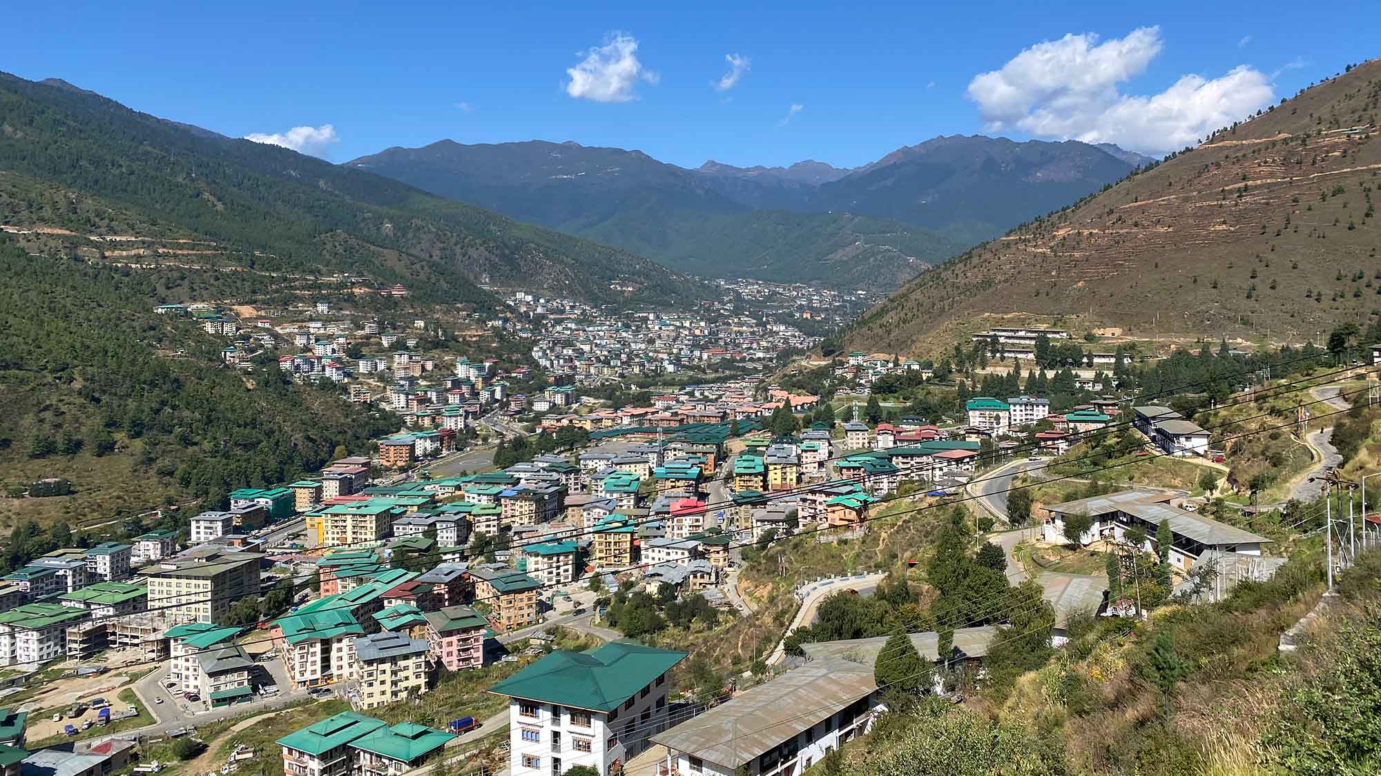 Photo of Thimphu showing buildings with multi-coloured roofs in the foreground and the mountains in the background.