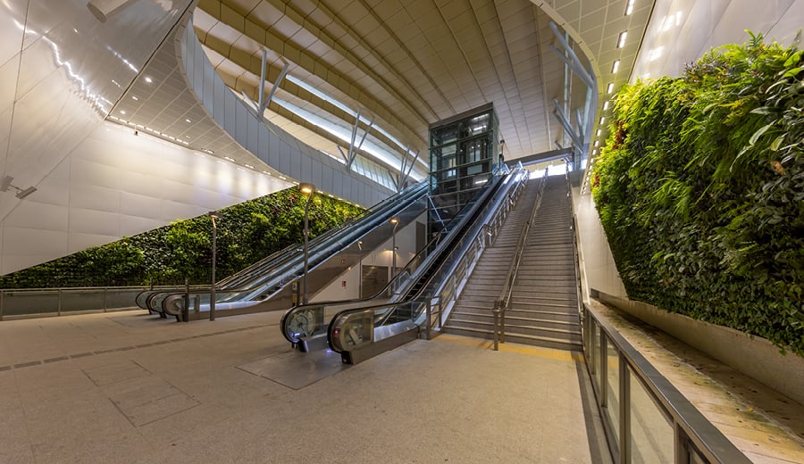 Underground walkway with escalators and stairs leading up to street level. Garden beds line the walkway. 