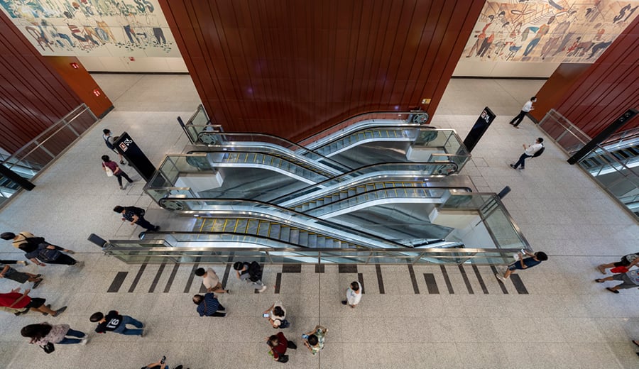 Inside a large modern underground rail station in Asia. Top down view of escalators and people moving through the space.
