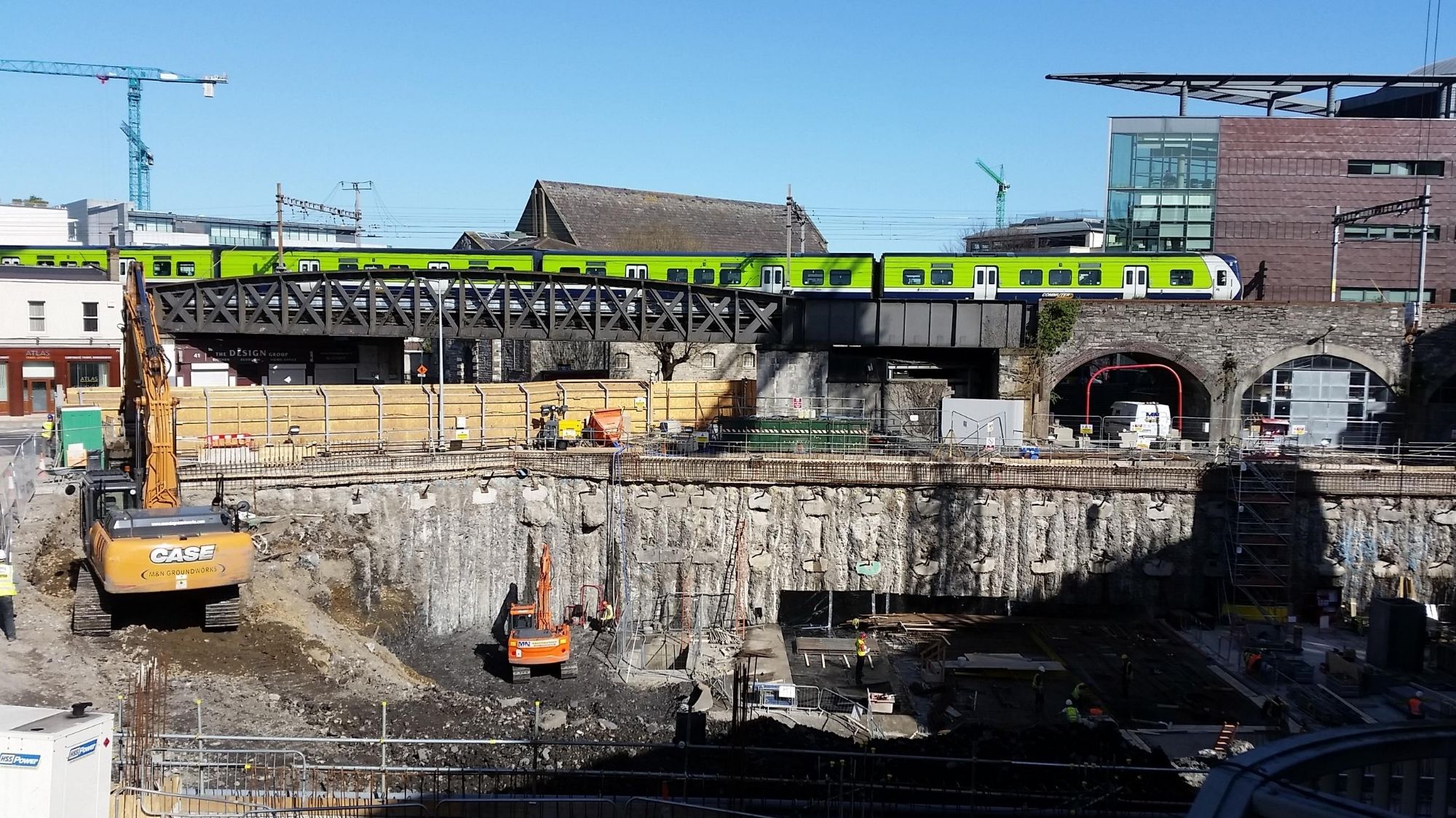 Construction photo of the site at Trinity Business School showing the excavation of the double-height basement alongside the railway line. A train is seen passing beside the construction site.