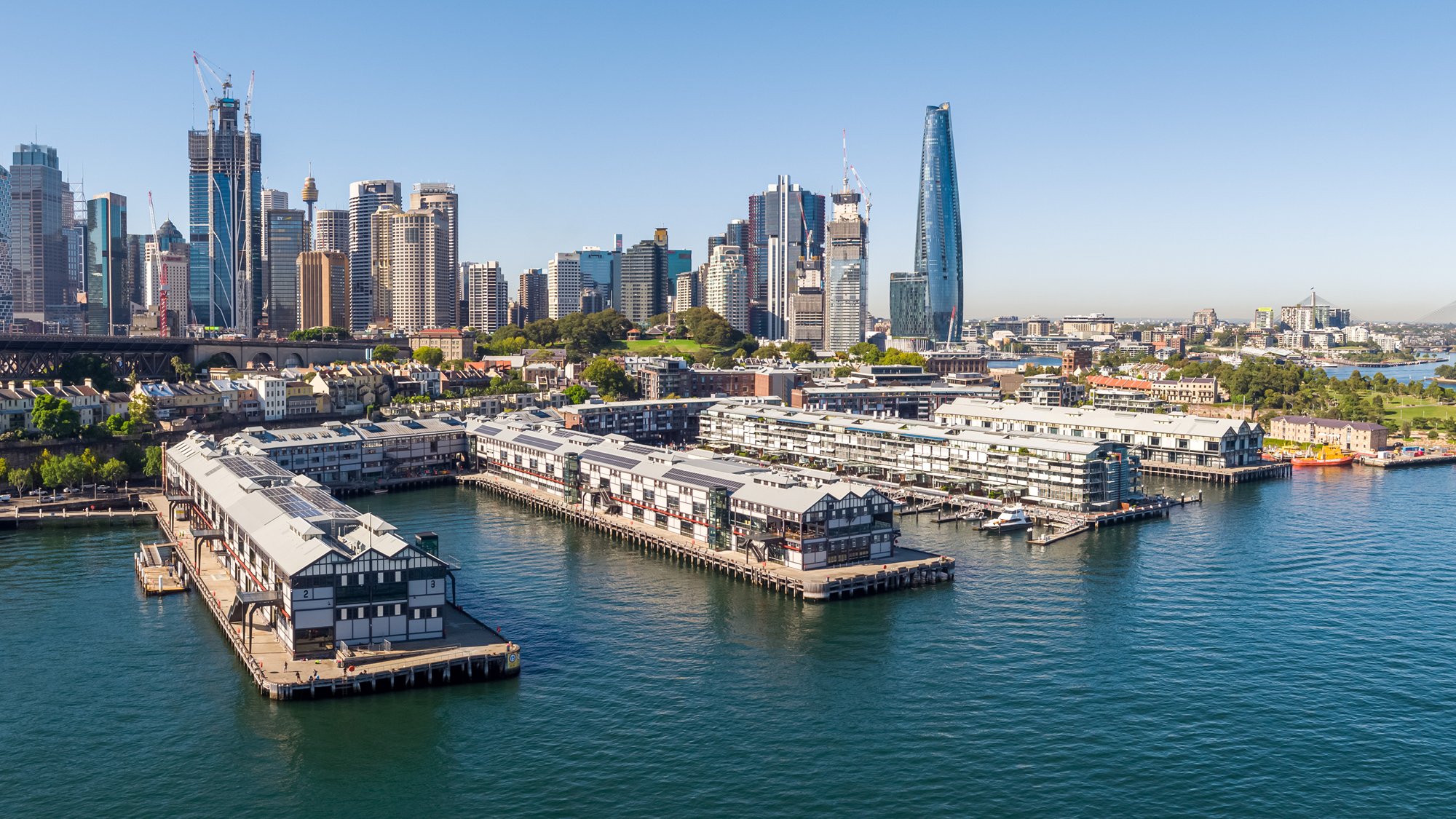 An aerial view of Walsh Bay piers on Sydney Harbour on a bright sunny day. City buildings are in the background. 