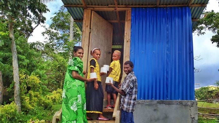 A Vanuatuan family holding toilet rolls while standing in front of a toilet building