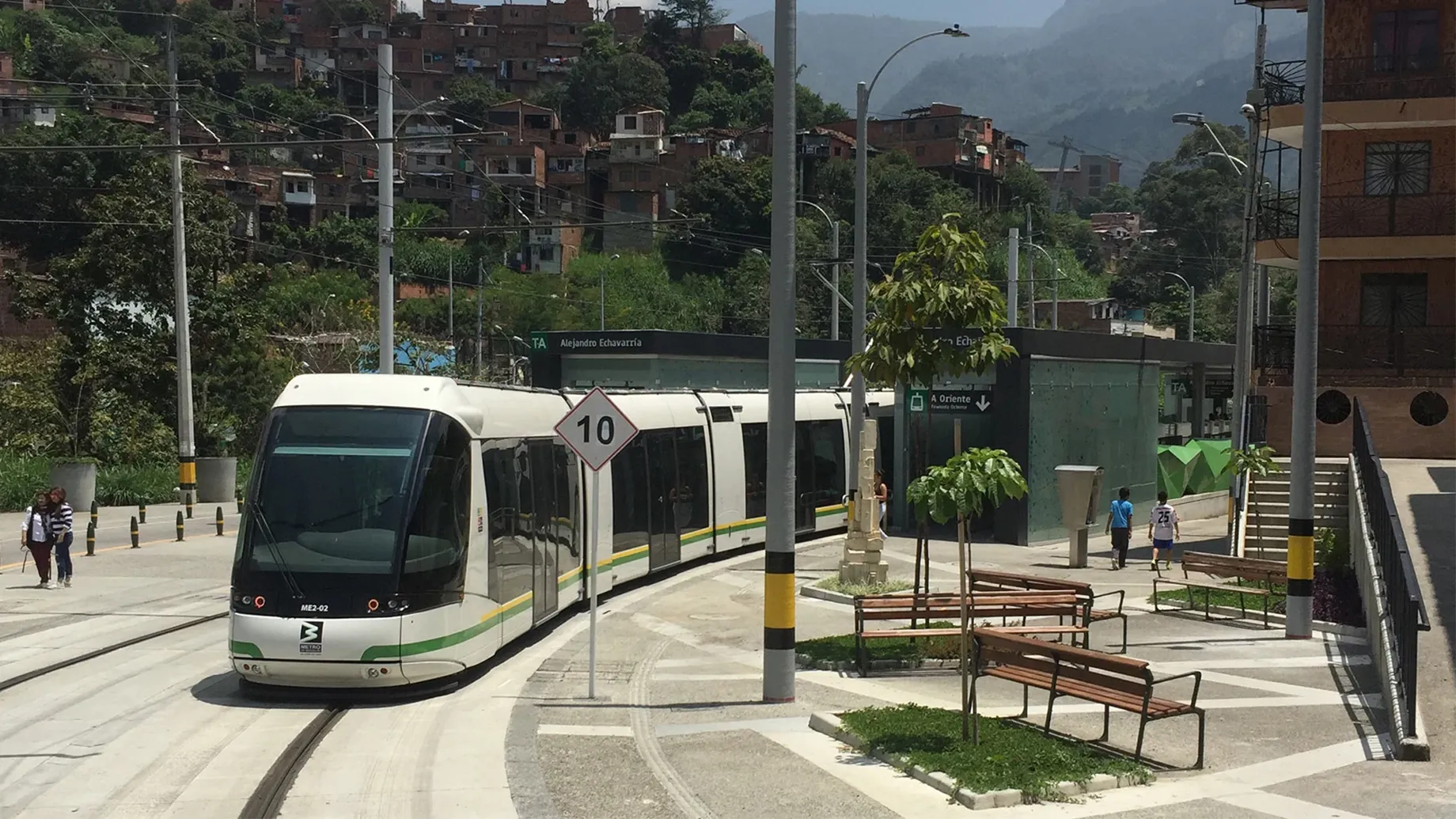 A train on street level tracks with park benches and houses on a hill in the distance