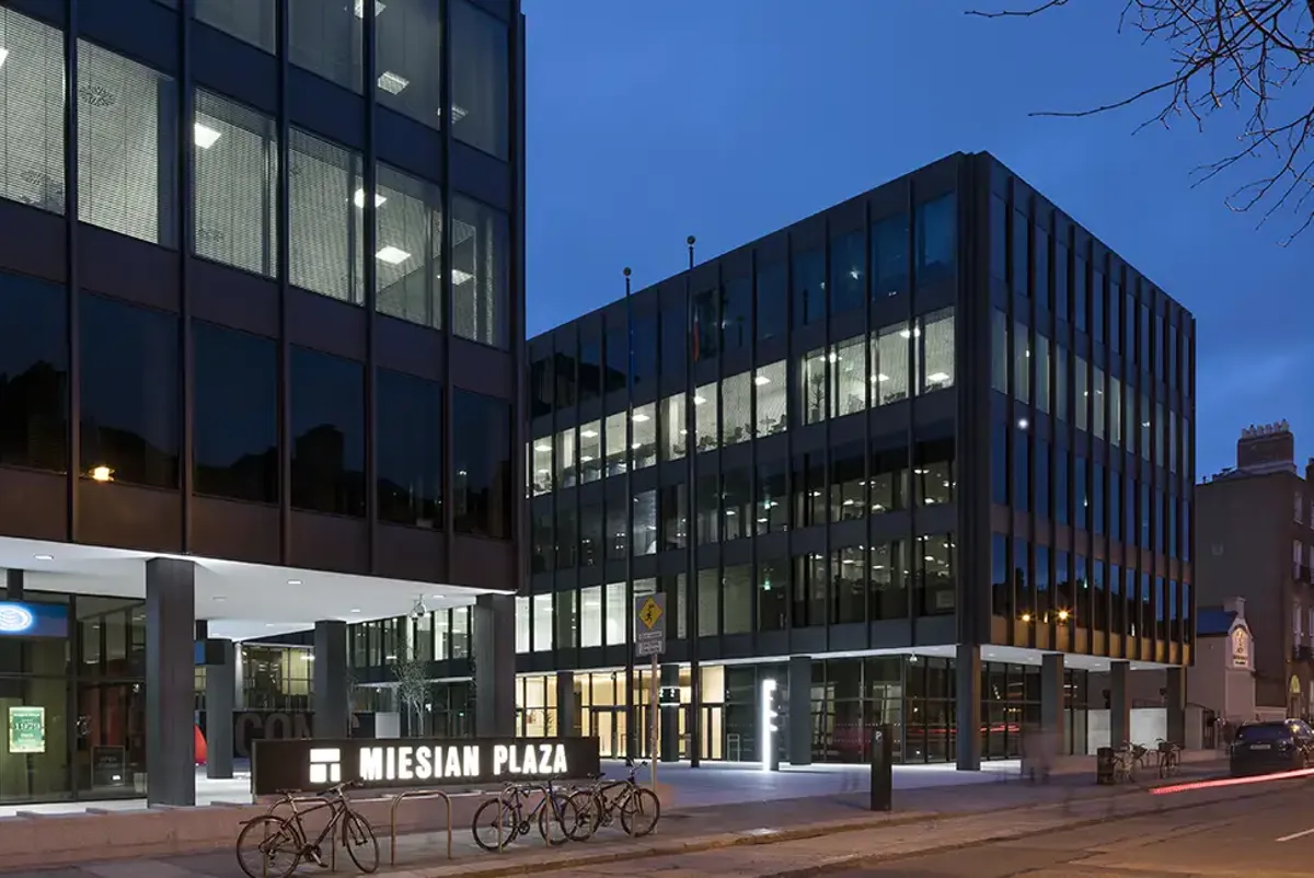 Night-time shot of Miesian Plaza buildings, with a sign lit up saying 'Miesian Plaza' and bicycles parked in front.